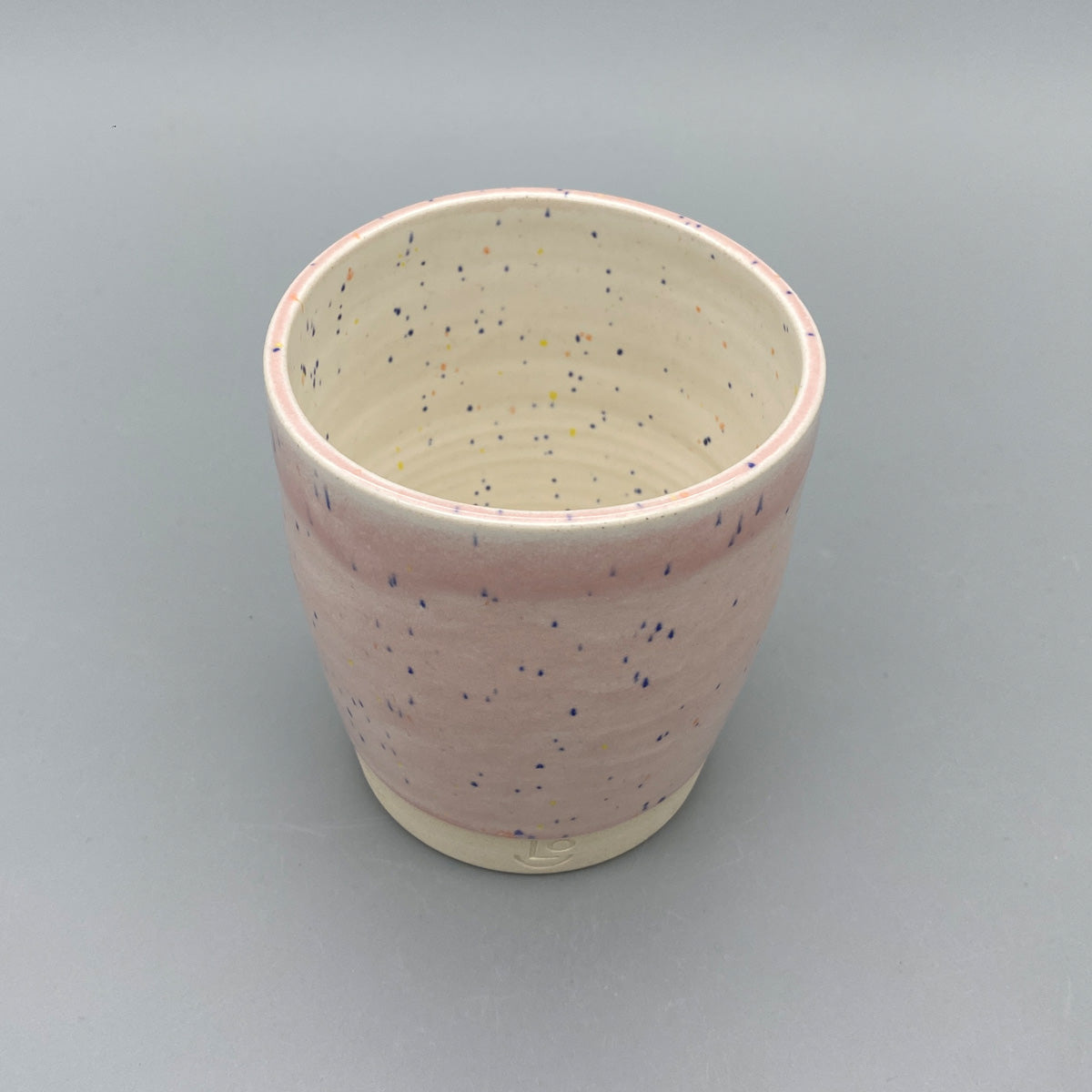 A pink ceramic cup/tumbler with blue speckles, placed on a plain grey background. The bottom portion is left unglazed, revealing the natural stoneware with the potter's mark "LO" (Liam Owen) inscribed.