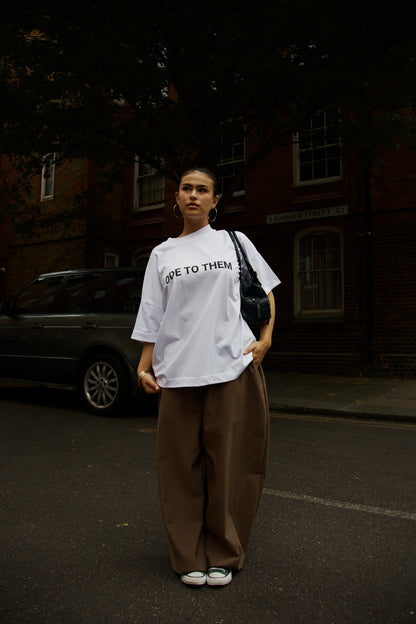 Woman wears Ode To Them baggy/oversized white graphic slogan T-shirt with Gargoyle loose fit trousers. She looks contemplative trainers and hoop earrings  