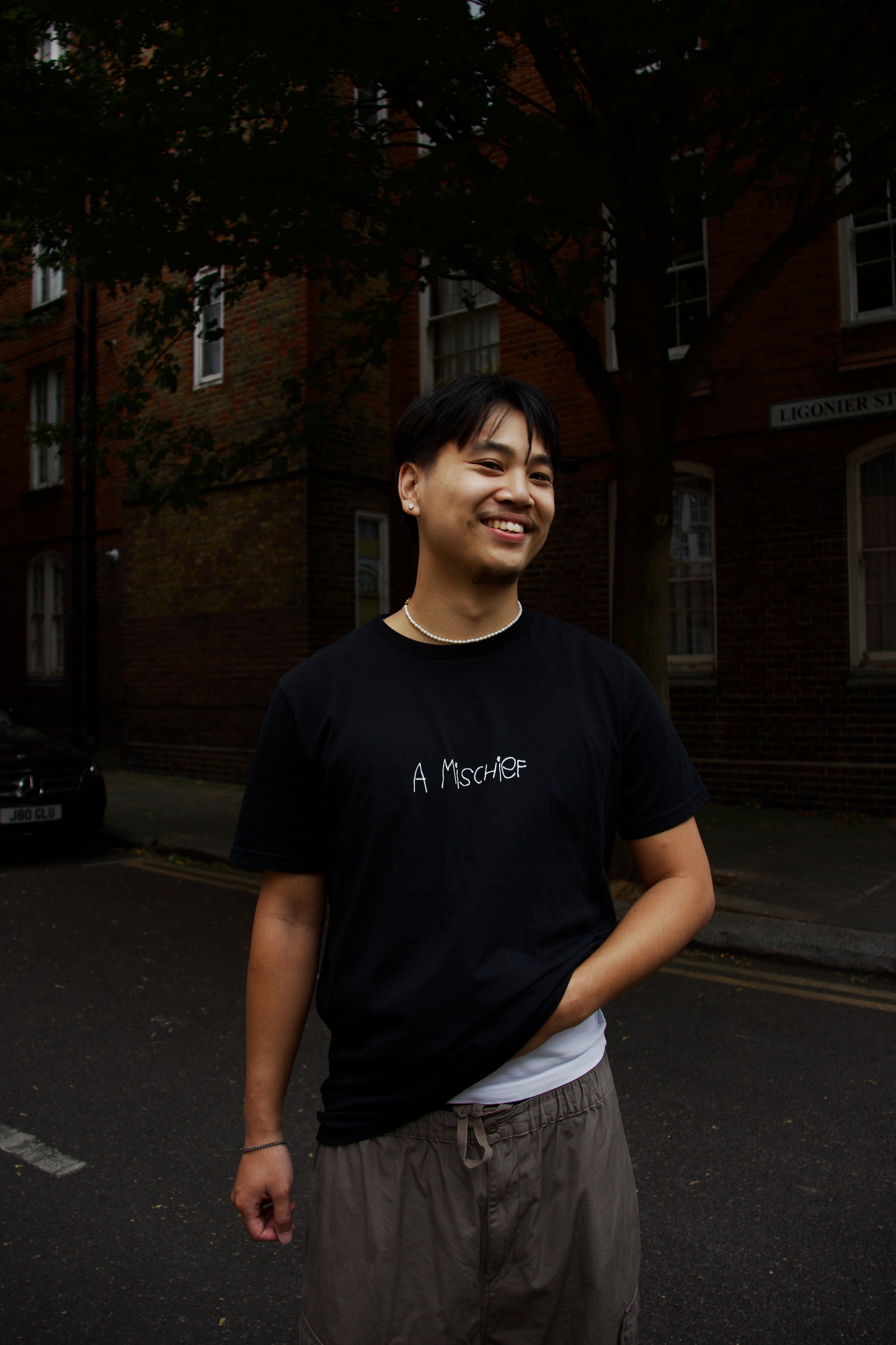 Man wears a mischief black baggy tee with loose trousers and is smiling cheekily 