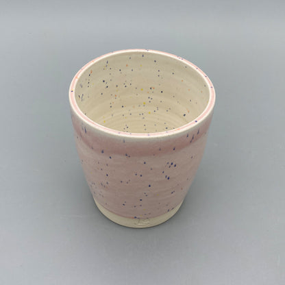 A pink ceramic cup/tumbler with blue speckles, placed on a plain grey background. The bottom portion is left unglazed, revealing the natural stoneware with the potter's mark "LO" (Liam Owen)  inscribed.