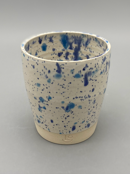 A white ceramic cup/tumbler with blue speckles, placed on a plain grey background. The bottom portion is left unglazed, revealing the natural stoneware with the potter's mark "LO" (Liam Owen)  inscribed.